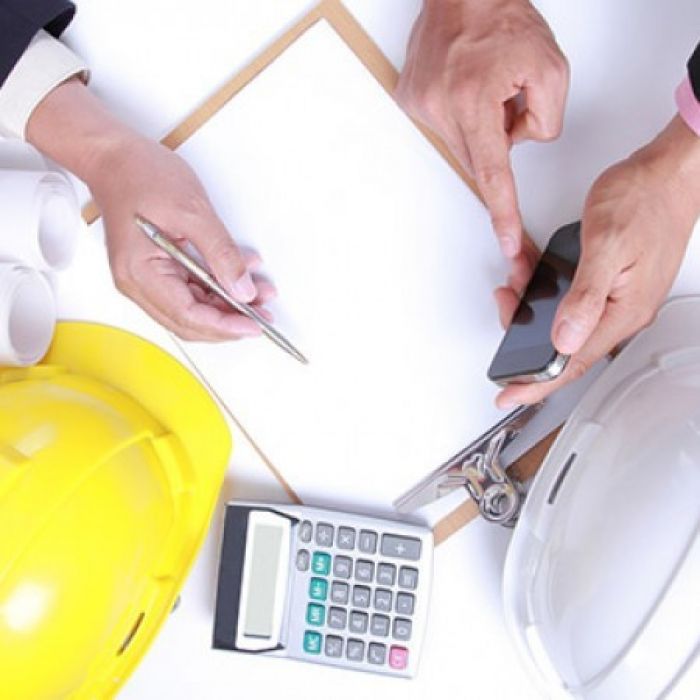 flatlay van een clipboard met papier, een rekenmachine, een witte en gele helm en twee paar handen met een pen en een telefoon